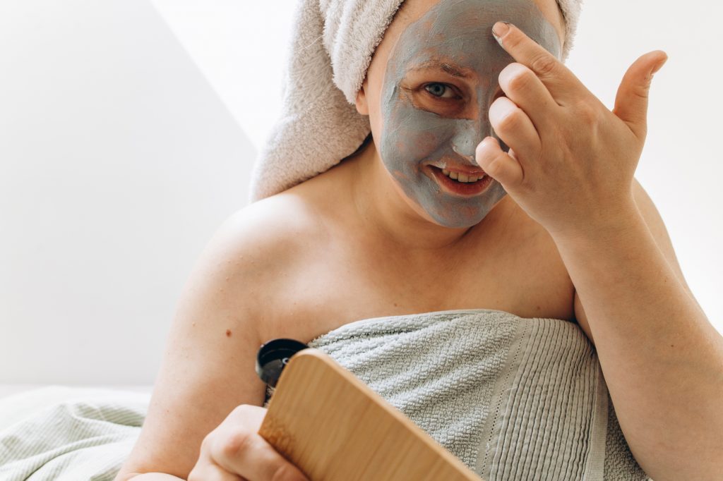 Happy plus size woman Applying Beauty Mask On Her Face with portable mirror wearing towel on hair At Home.